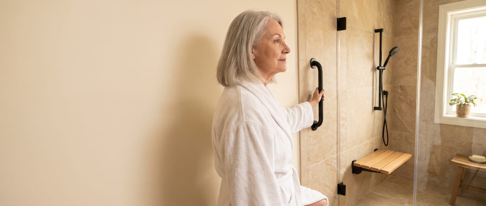 An older woman steps into a level-entry shower, one hand on a matte black grab bar beside a fold-down bench